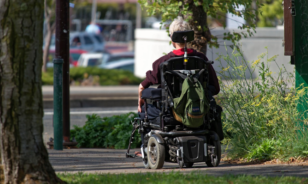 En som kör en permobil, elektrisk rullstol. Bilden är tagen så man ser den bakifrån, och en ryggsäck hänger baktill. På en gångväg med träd och växter runtom. Ser ut som en stadsbild.
