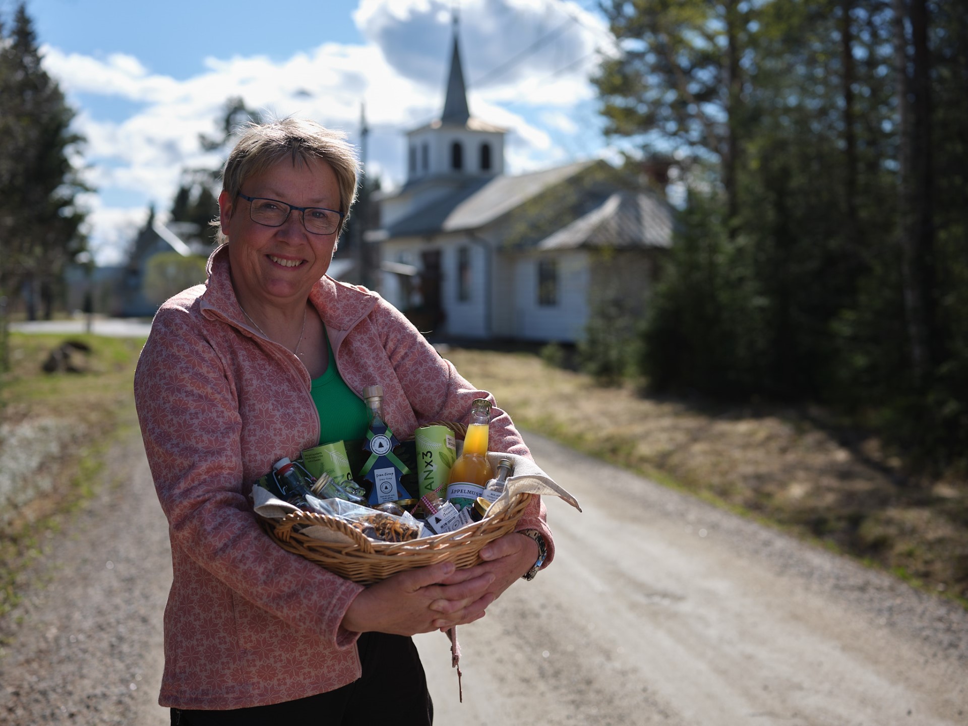 Eva med en korg i famnen fylld med lokala produkter på byvägen. Det gamla kapellet i bakgrunden.