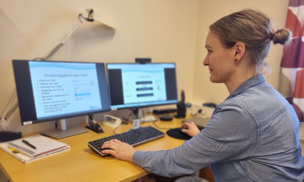 Erica sitter framför skrivbordet med två skärmar. Hon har blå skjorta och uppsatt hår.