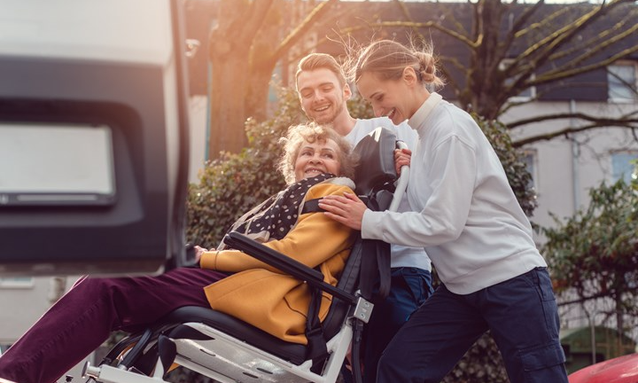 patient på väg in i en taxi med hiss. två personer håller i på var sin sida.