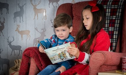 Flicka läser bok för yngre pojke. De sitter i en röd fåtölj. Vit tapet med hjortar.