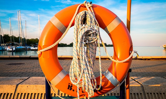 orange livboj som hänger vid en kajkant. Segelbåtar ligger förtöjda i bakgrunden. Bakom en udde med träd.Blå himmel och horisont.