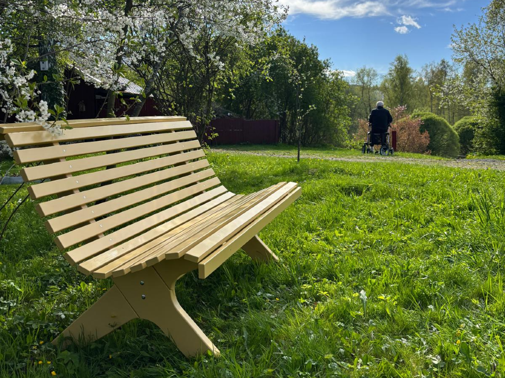 Gul bänk i gräset vid en promenadväg. Lummigt och grönt med blommande trädgren hängande över bänken. I bakgrunden en person gående med rullator