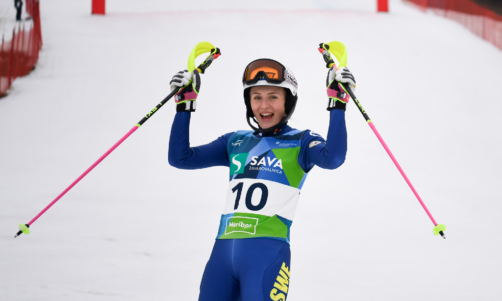 Ebba står längst ner i slalombacken i målhanget och sträcker upp armar och stavar i skyn. Hon har skiddräkt i svenks afärger och hjälm med glasögon uppe på. Grön nummerlapp med siffran 10.
