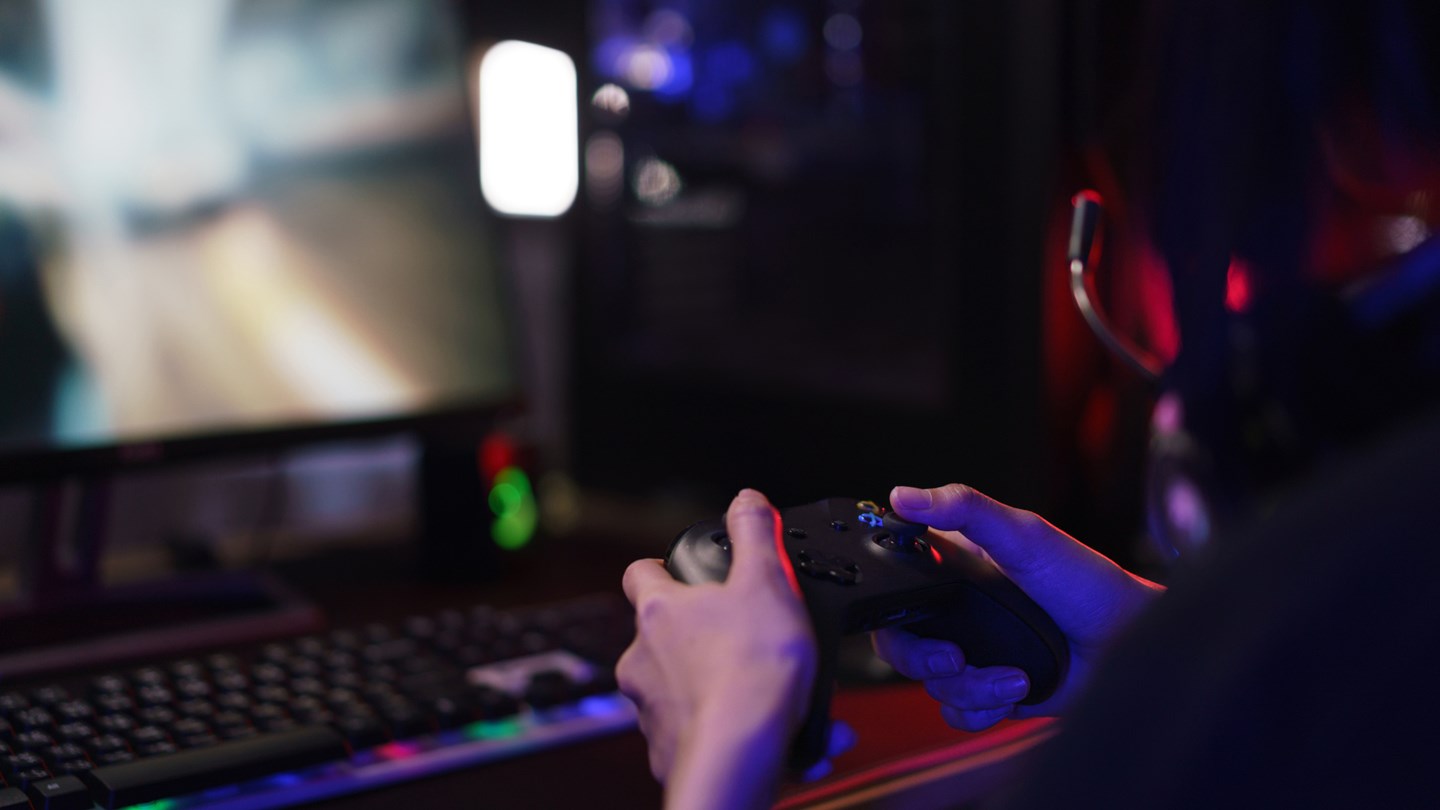 En som håller i en spelkontroll och spelar ett dataspel. Det är mörkt i rummet, förutom ljuset från skärmen.