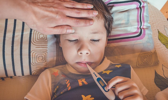 barn ligger i en säng och håller en febertermometer i munnen. En vuxen känner med handen på barnets panna.
