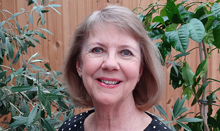 En glad Eva Ullerud framför gröna växter. Hon har ljusbrunt hår nästan ner till axlarna.