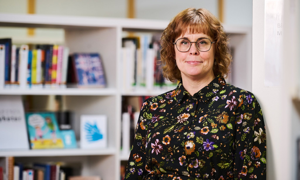 Desirée Stensdotter i blus med blommor på svart botten. Hon har ljusbrunt lite lockigt hår till axlarna. Hon bär glasögon. Hon står framför vit hylla med böcker.