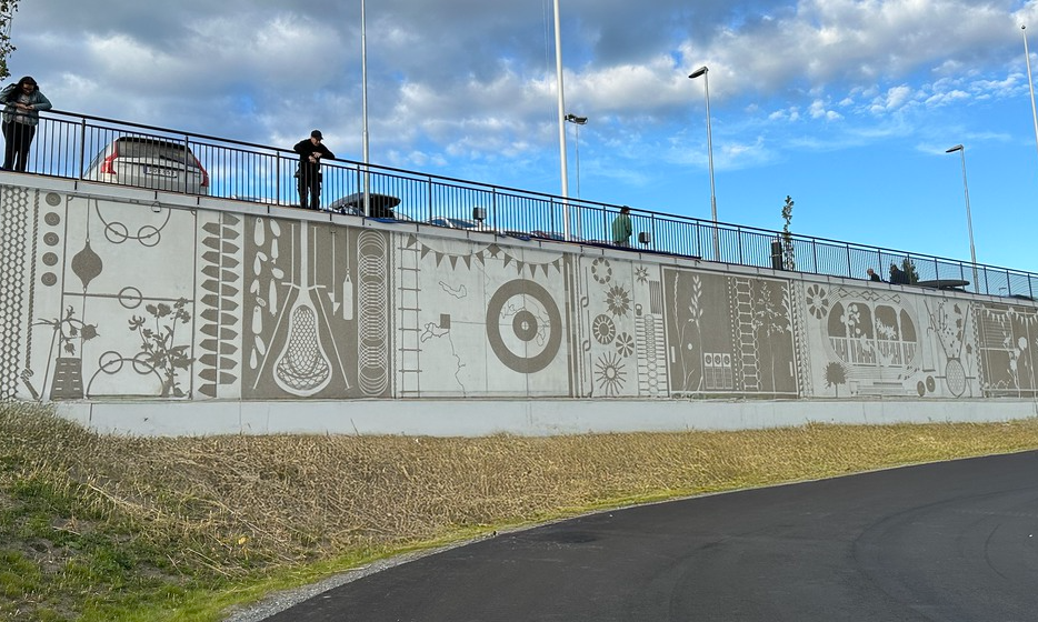Muralkonstverk i grafisk betong med grön relief. De olika delarna föreställer lek, sport och växter.