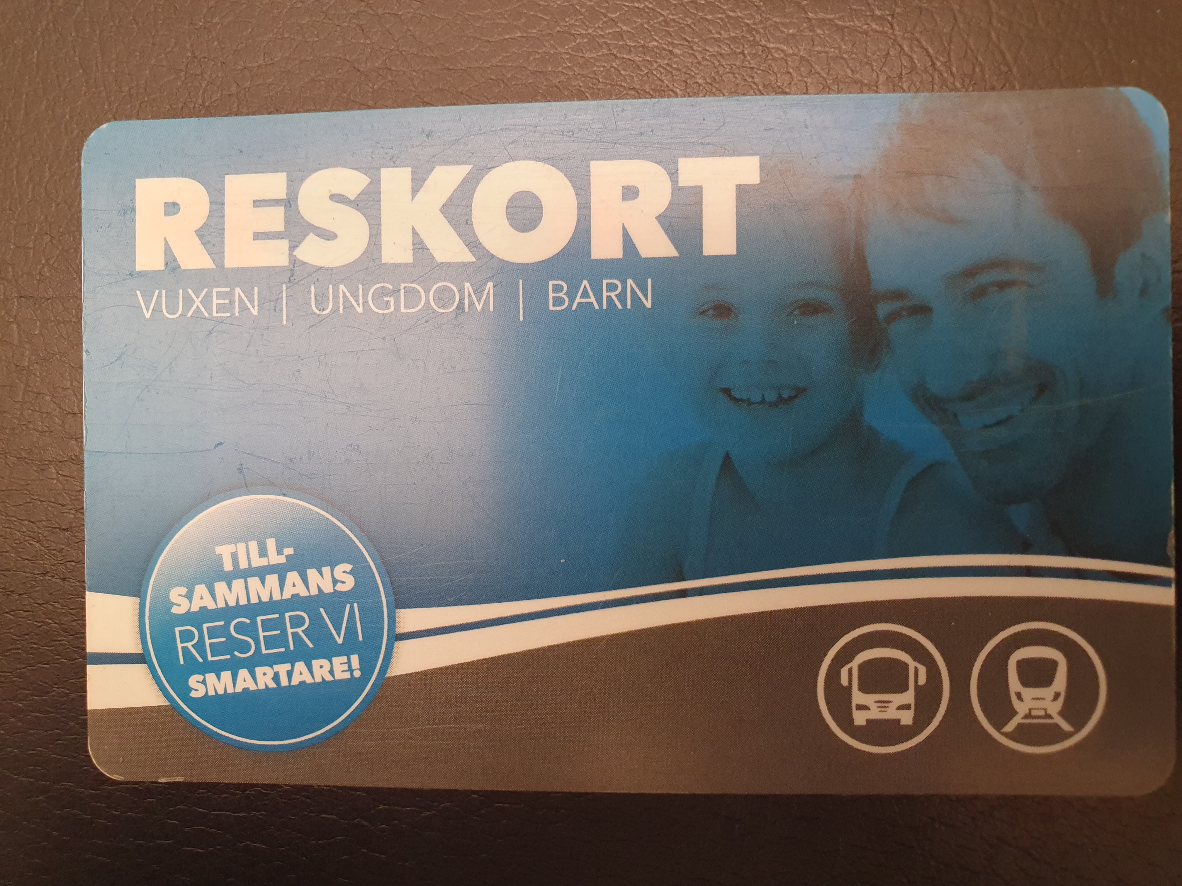 Reskort i blått och svart, med vita ränder mellan det blå och svarta. En bild på en vuxen och ett barn på det blåa. Symboler för buss och tåg på det svarta. På en blå cirkel står det Tillsammans reser vi smartare. Det står Reskort med vit text överst i mitten..