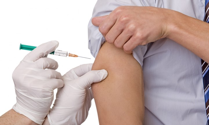 Närbild på överarm. Personen håller i den upprullade skjortärmen. Personen får en vaccination i överarmen. Handskklädda händer håller i sprutan.