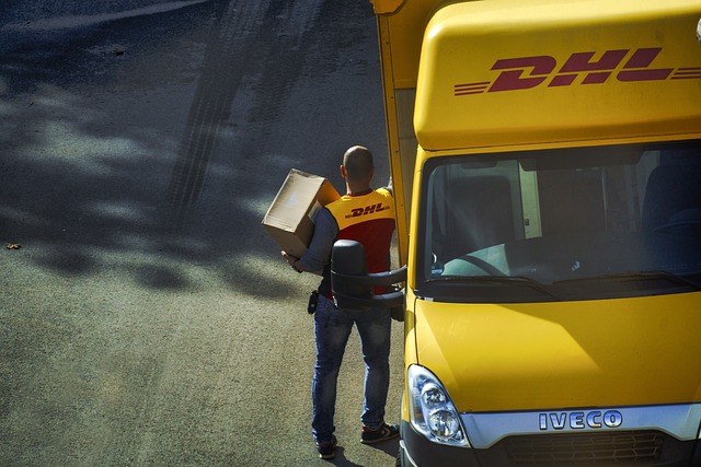 Fronten på en gul budbil. En person har tagit ut ett paket från budbilen. Det står DHL med röda bokstäver på budbilen och personens rygg.