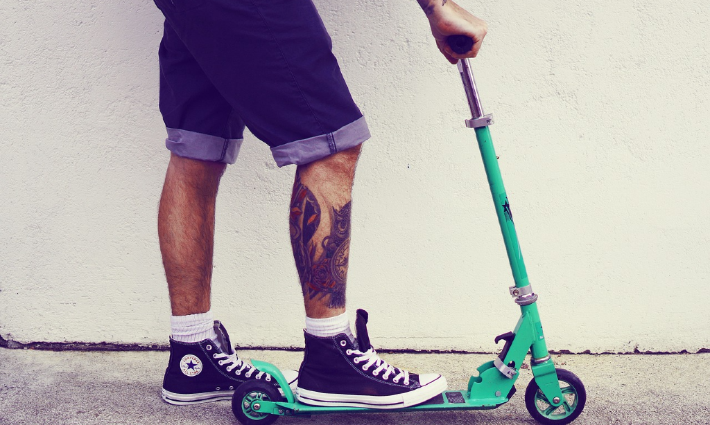 Person i jeansshorts och tatuerat höger ben står på en grön sparkcykel.