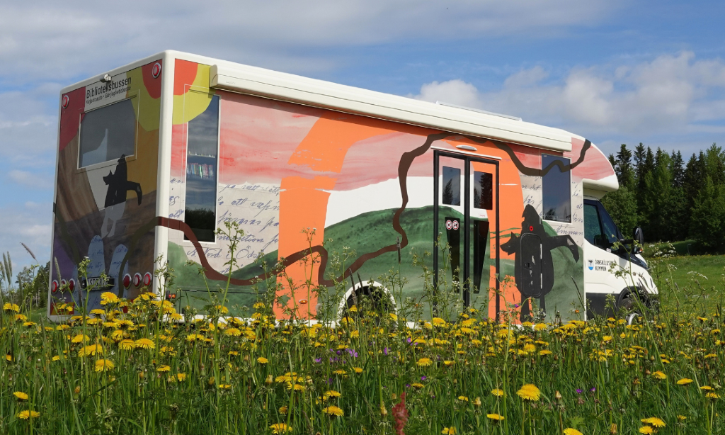 Bokbussen med naturmålning i grönt och orange. Den står vid en sommargrön äng och gula blommor.