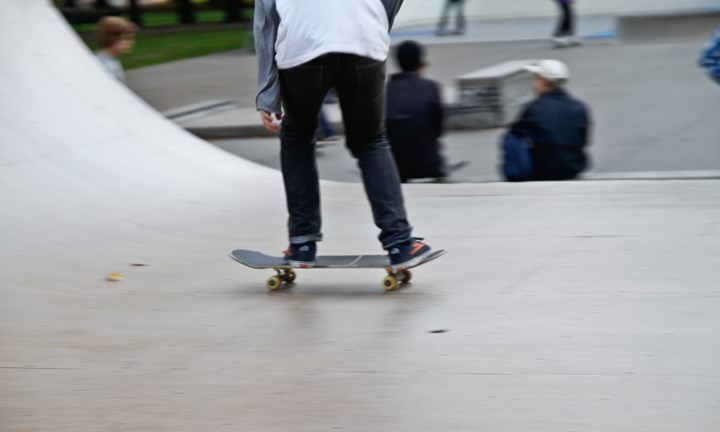 En person med mörka jeans står på skateboard på en skateramp. Överkroppen syns inte. Nedanför skaterampen står lite folk.