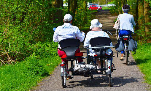 Ledsagare skjutsar en äldre person på parcykel på gångväg i en lummig park.