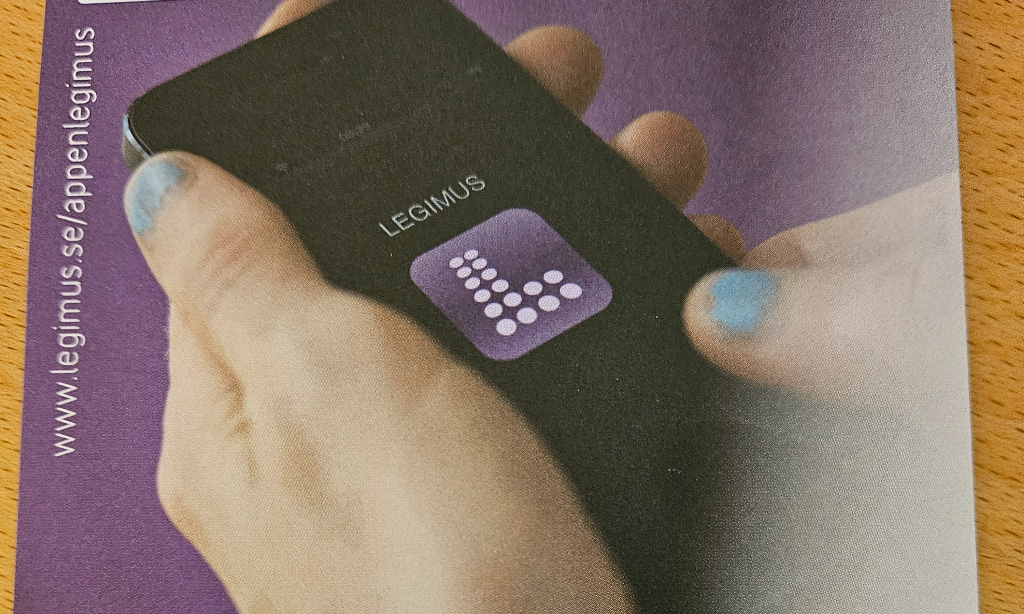 närbild på fingrar (blått nagellack)som håller i en mobil. På skärmen syns loggan för Legimus appen. En lila kvadrat med L format av vita punkter. Det står LEGIMUS ovanför i vitt.