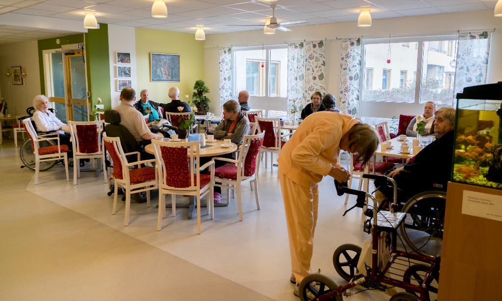 Äldre sitter samlade i en ljus samlingslokal på äldreboende.
